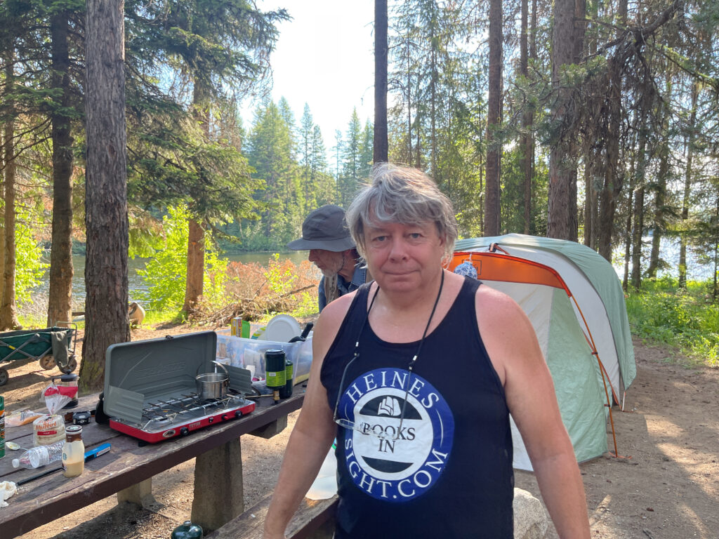 Matthew Heines standing at his forested campsite in the Northern Cascade Mountains wearing a black tank top with Heines Books in Sight logo, with tent, picnic table, and lake in the background during the 2021 Bigfoot investigation with Igor Burtsev.